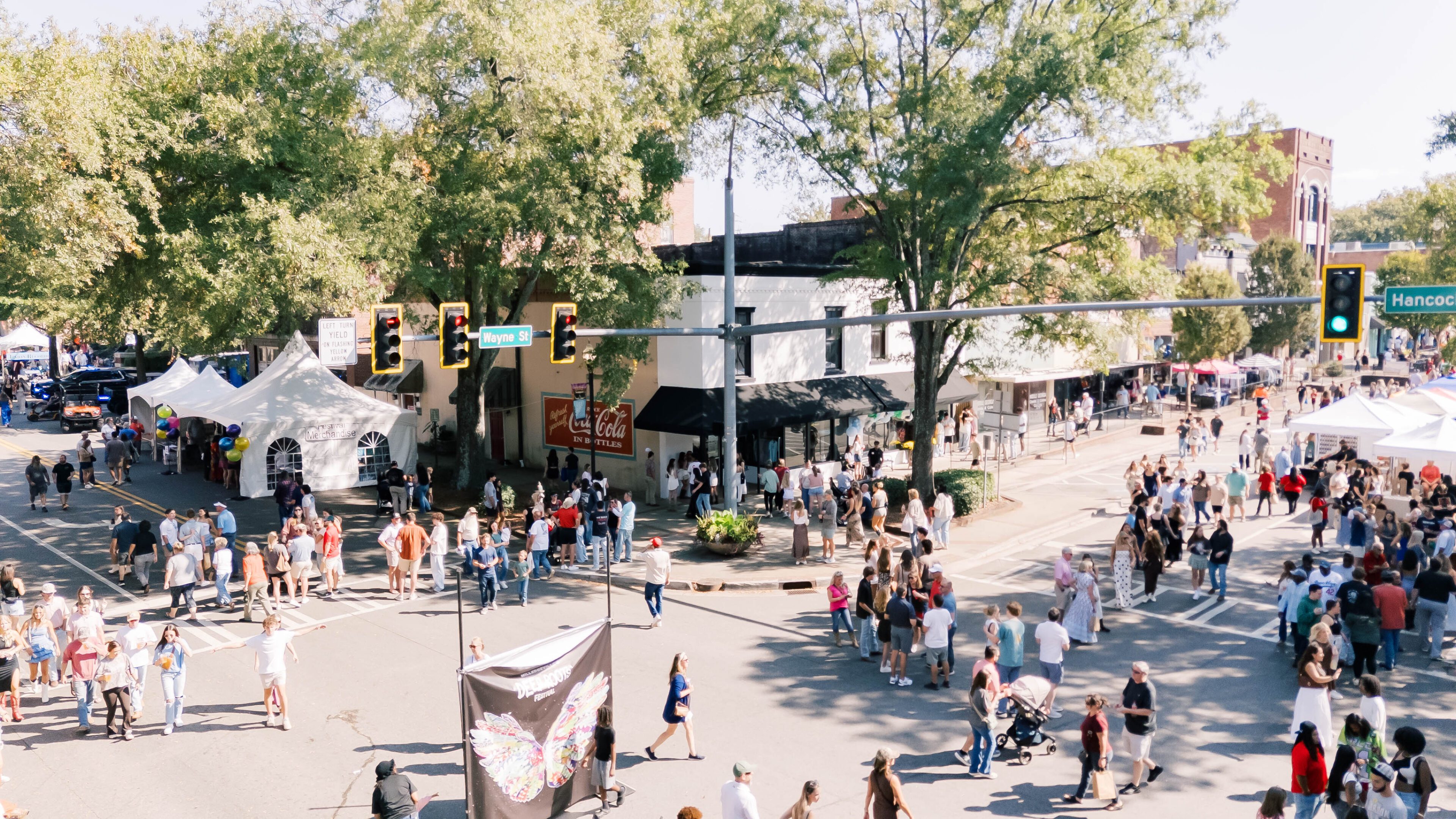 A lively outdoor market with people shopping under large trees.